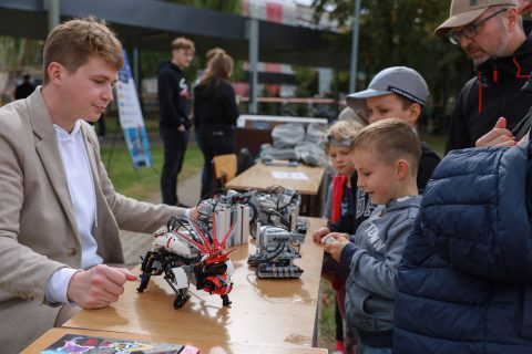 two boys, a girl and a man are looking at the robots made of building blocks standing on a table, the constructions are presented by a BUT student